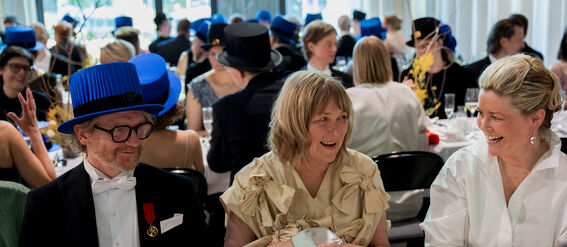 People at a formal event sitting at a table, many wearing blue hats. A woman holds a silver clutch.