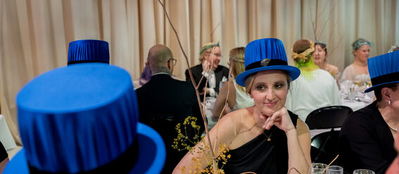 People seated at a banquet wearing blue hats with black bands, elegant table setting with flowers and drinks.