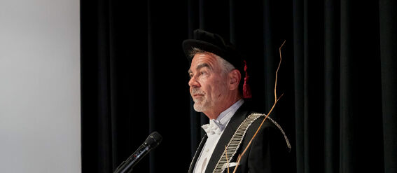A person in formal attire standing at a microphone with black curtains in the background.