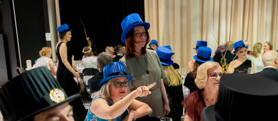 A formal dinner gathering with guests wearing blue and black top hats, seated at a table set with wine glasses and cutlery.