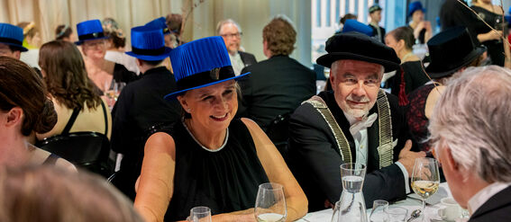 People seated at a formal event with blue and black hats, holding wine glasses, white tablecloths.