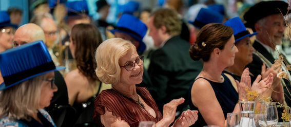 People seated at a formal event, clapping. Blue hats, elegant clothing, table settings with glasses and plates.