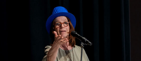 Person in grey dress and blue hat speaking into a microphone, standing at a podium with papers.