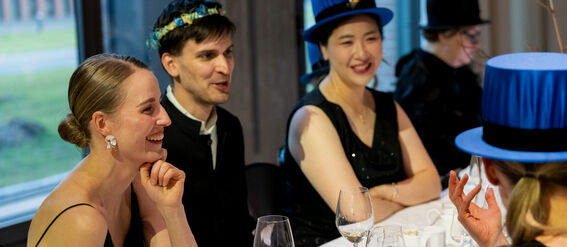Group of people at a formal event, seated at a table with wine glasses. Some are wearing blue hats and black outfits.