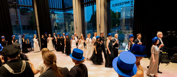 A group of people in formal attire dancing in a hall with large windows, some wearing blue top hats.