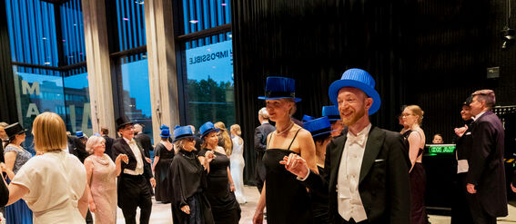 People dancing at an indoor formal event. Many are wearing blue hats and elegant attire. Large windows reveal evening outside.