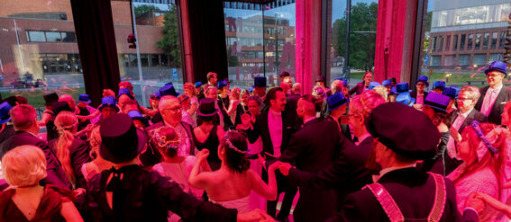 A formal dance event with people in elegant dresses and suits dancing in a circle, seen in a brightly lit room with large windows.