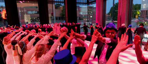 A group of people dressed formally participate in a dance under pink lights, extending their arms to form arches.