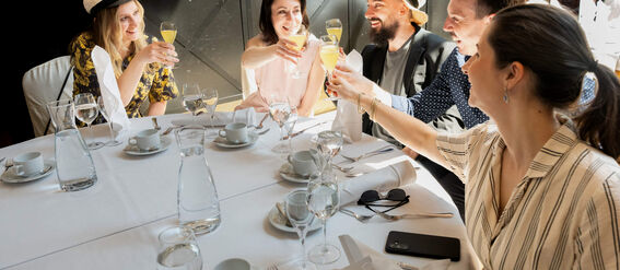 Five people sitting at a dining table, raising their glasses in a toast. The table is set with teacups, glasses, and utensils.