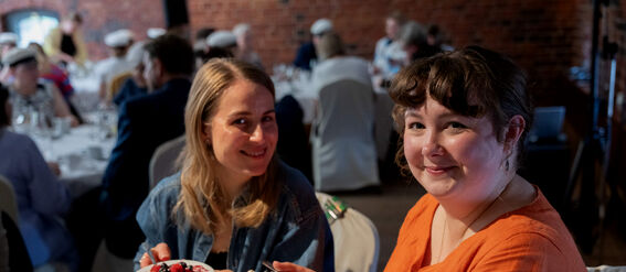 Two people holding plates of dessert at a formal gathering, seated at a table with a white tablecloth.