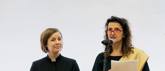 Two women at a podium; one holds notes, the other a folder. One wears a black robe and the other a gown with a gold sash.