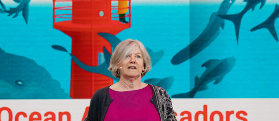 A person in a magenta dress and grey cardigan stands before an ocean-themed background with text.