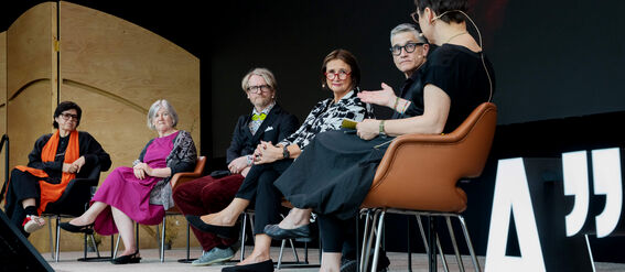 Six people sit on stage in a discussion panel. A large quotation mark symbol is visible on the right.