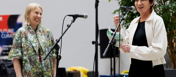 Kaksi naista seisoo mikrofonien lähellä, toisella kukkakuviollinen puku, toisella musta mekko ja valkoinen takki.