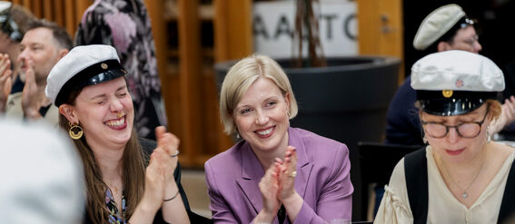 Ryhmä ihmisiä juhla-asuissa taputtamassa. Nainen violetissa takissa, valkoiset lakit. Shamppanjalasit ja lautaset pöydällä.