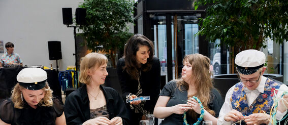 Ihmisiä istuu pöydässä tekemässä käsitöitä langalla. Pöydällä on samppanjalaseja, leivonnaisia ja värikkäitä materiaaleja.