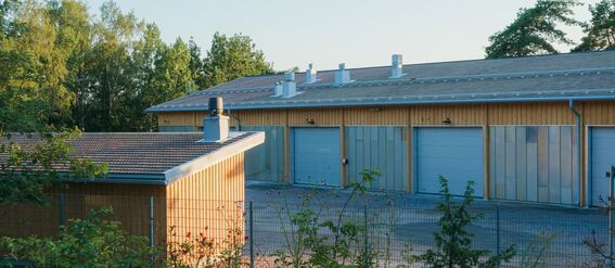 Wooden, storage buildings with grey roofs behind a fence and vegetation in the foreground.