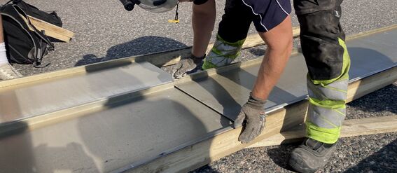 A worker in protective gear is looking at aluminum panels on wooden frames outdoors. Graffiti and vehicles are visible in the background.