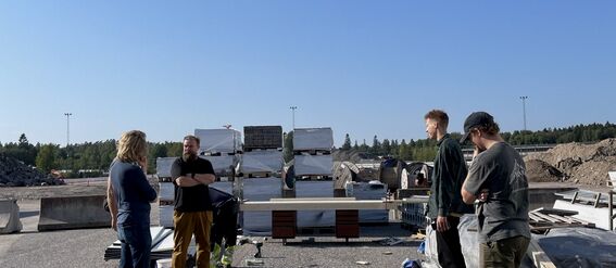 Several people standing beside construction materials on a sunny day in an industrial area with a clear blue sky.