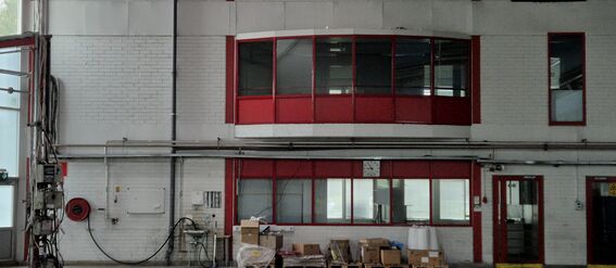 Industrial warehouse interior with white walls and red-framed windows and doors. Boxes and equipment on the floor.