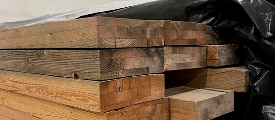 Large stack of glulam wrapped in plastic on one side, with industrial pipes visible in the background.