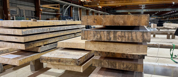 Stacks of large glulam planks with markings in a spacious industrial warehouse with high ceilings and various pipes.