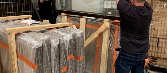 Two men are handling a large glass window in an industrial setting. Wrapped items are stacked nearby.