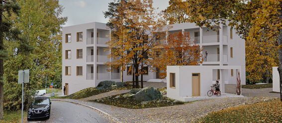 A white apartment building surrounded by autumn trees and fallen leaves. A person rides a bicycle near a small white structure.