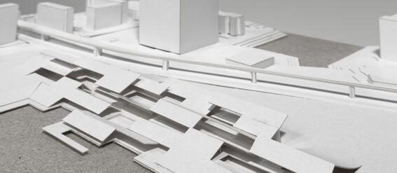 A white architectural model with various rectangular structures and a tall building, displayed on a grey surface.