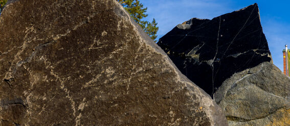 Lähikuva kahdesta suuresta kivestä, toinen tummanharmaa ja toinen musta, taustalla puita ja osittain pilvinen sininen taivas.