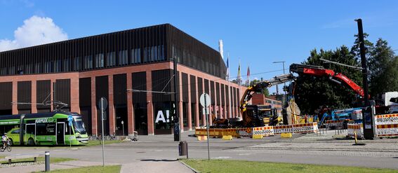 Punainen ja keltainen rakennustyömaa, vihreä raitiovaunu modernin tiilirakennuksen edessä kirkkaan sinisen taivaan alla.