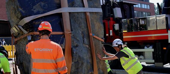 Työntekijät asentavat suurta, hihnoilla kiinnitettyä kiveä nosturin avulla. He ovat ulkona aurinkoisena päivänä.