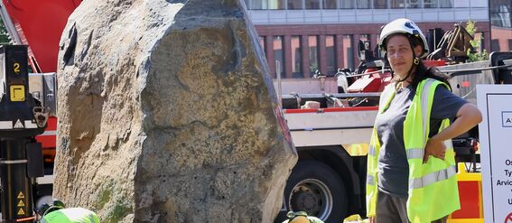 Rakennustyöntekijät huomioliiveissä ja kypärissä työskentelevät suuren kiven lähellä, taustalla raskaita laitteita.