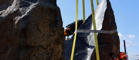 Oranssi rakennustyöntekijä nostaa kahta isoa kiveä ketjuilla ja hihnilla sinisen taivaan alla.