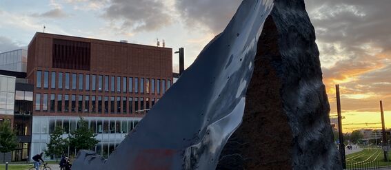 Suuri veistos etualalla, taustalla moderneja rakennuksia ja pyöräilijöitä, pilvinen taivas auringonlaskun aikaan.