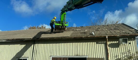 A worker in safety gear on an old tiled roof under a blue sky.