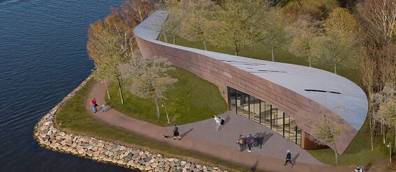 A modern, curved building with a silver roof by a lakeside. People walk on a pathway surrounded by trees.