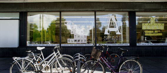 Several bicycles are parked in front of a building with large windows displaying signs about summer opening.