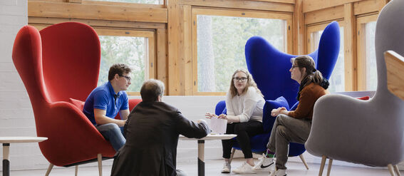 Four people having a discussion in a modern room with large windows. They sit on colourful chairs and cushions around small tables.