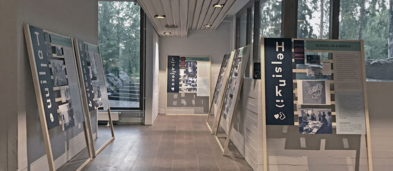 Exhibition room with large posters on wooden stands. Posters display city names like Helsinki, Tallinn, and Ljubljana.