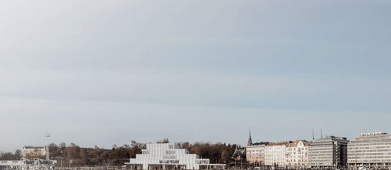 A waterfront view of a city with modern buildings and a stepped glass structure, reflecting in the calm water.