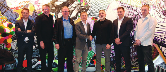 Six men in formal attire stand in front of a colourful abstract mural. Two men in the centre are shaking hands.