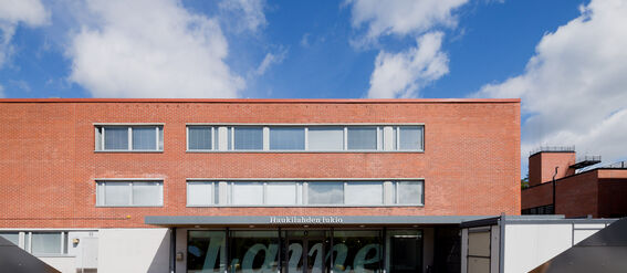A red-brick building with large windows and a sign reading 'Haukilahden lukio'. The sky is blue with scattered clouds.