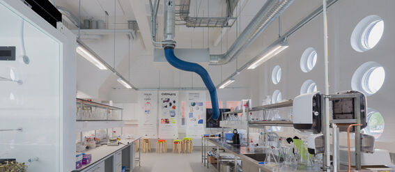 A modern laboratory with stainless steel benches, circular windows, and a blue ventilation pipe. Posters and stools are visible.