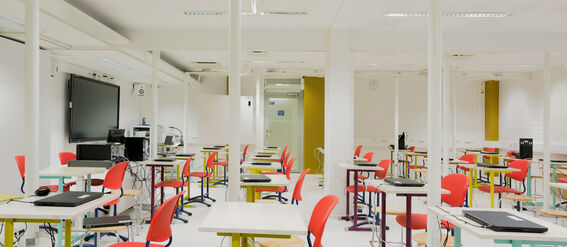 A modern classroom with red chairs, white tables, and computers. A large screen is mounted on the wall.