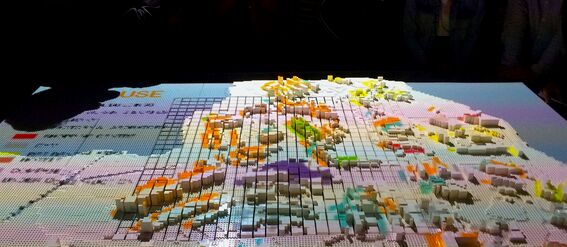 A 3D map with colourful blocks on a table, surrounded by people observing it.