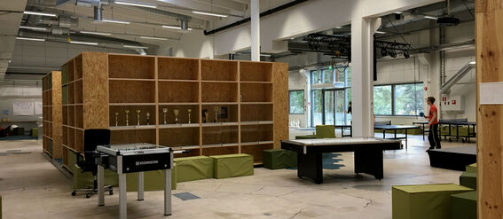 Spacious room with wooden shelves, table football, green seating, and people playing table tennis near large windows.