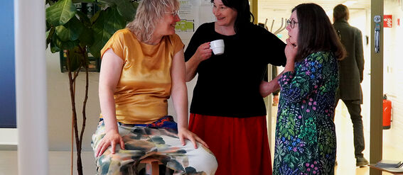 Three adults chatting indoors; one seated, two standing with a coffee cup, in colourful clothes by a tall plant.