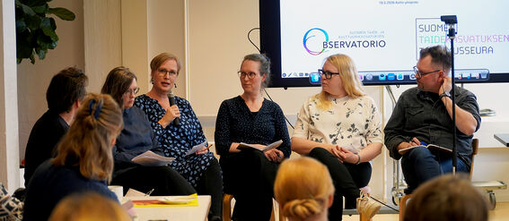 Panel of adults discussing art education in a classroom, audience seated, presentation slide on large screen.