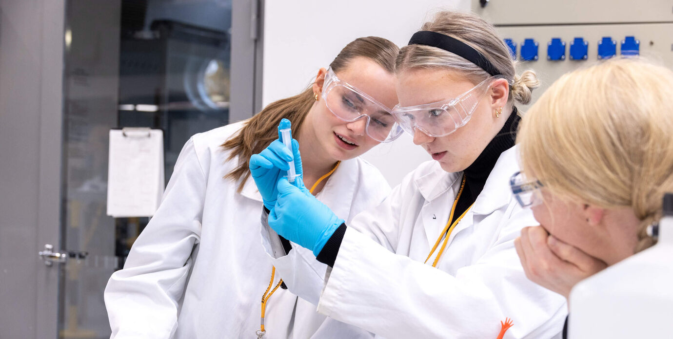 Three young womeen in lab coats observe a blue liquid in a test tube. They are inside a modern laboratory.